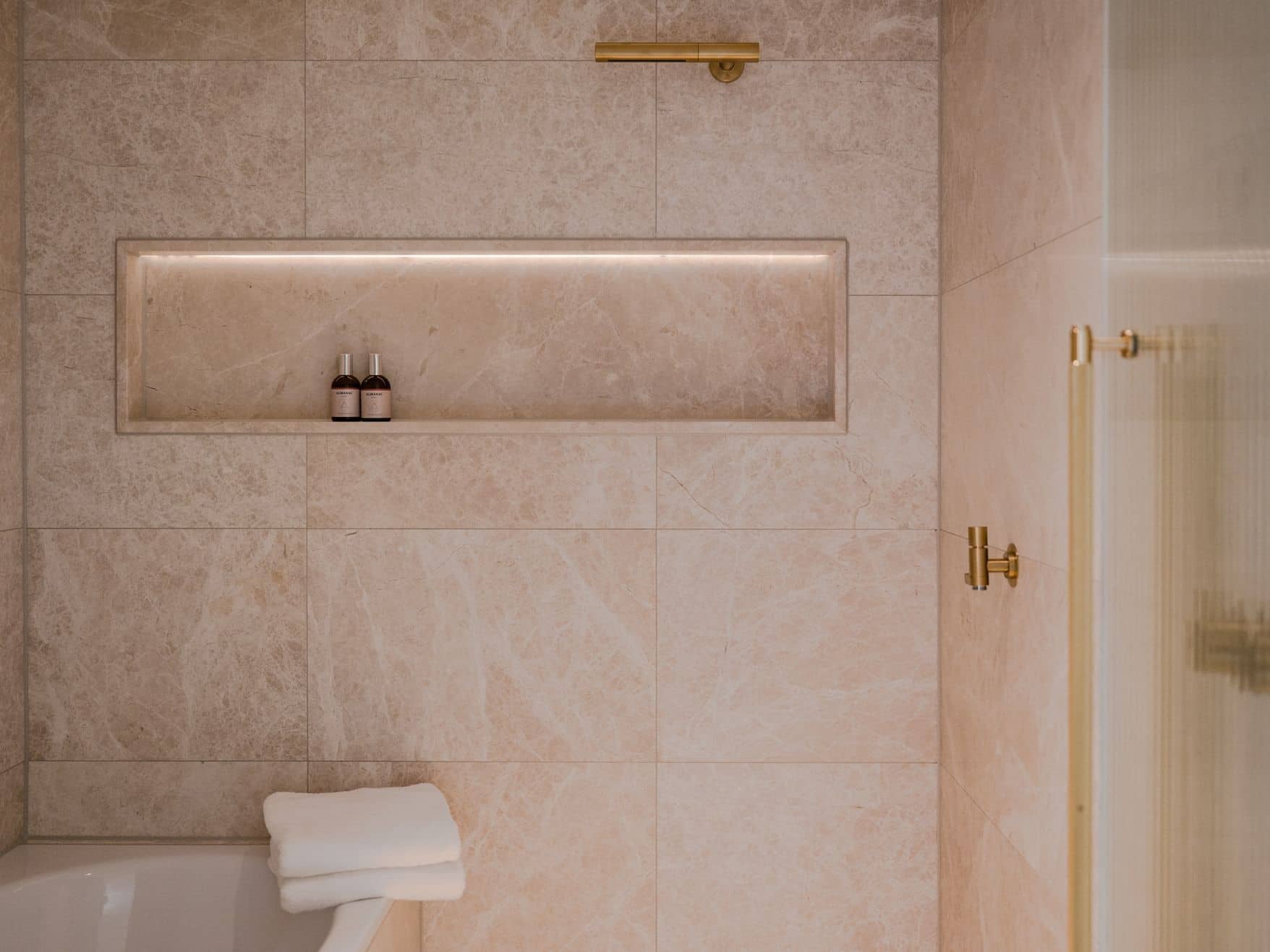 A modern bathroom in the Almanac Room features beige marble tiles, a built-in shelf with two small bottles, a folded white towel on the tub, and elegant gold fixtures.