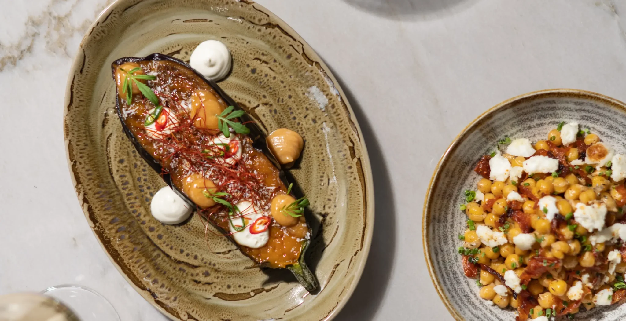 A roasted eggplant half garnished with sauce, herbs, and cream sits on a ceramic plate next to a bowl of chickpea salad with feta and herbs.