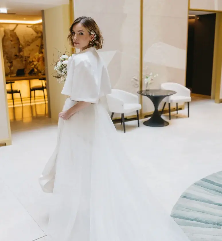 A woman in a white wedding dress holding a bouquet stands in a modern, light-filled room with white chairs and a round black table in the background.