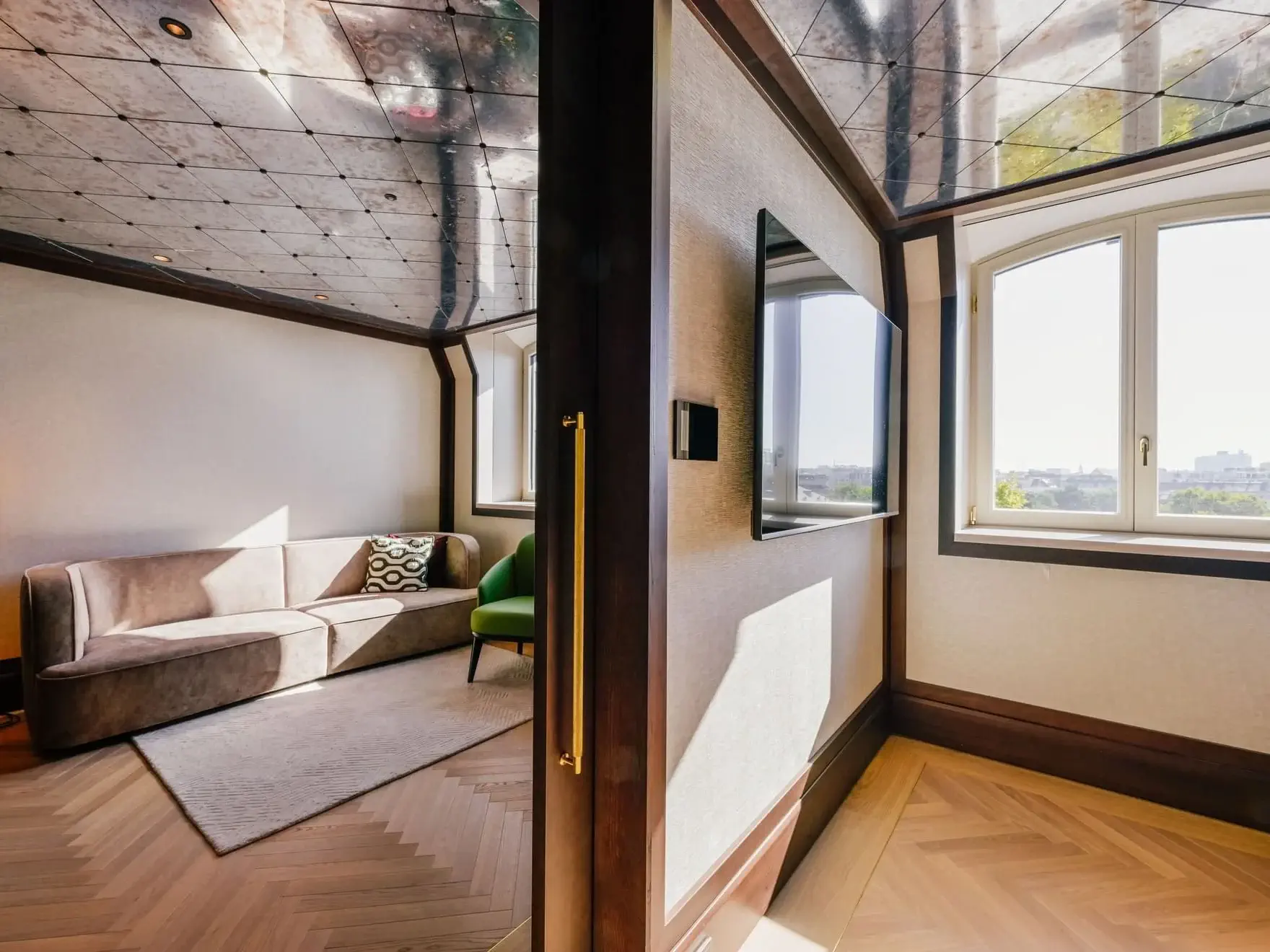 A corner of the Almanac Park Suite features a beige sofa, green chair, wall-mounted TV, large window, and a reflective ceiling that reveals striking structural elements.