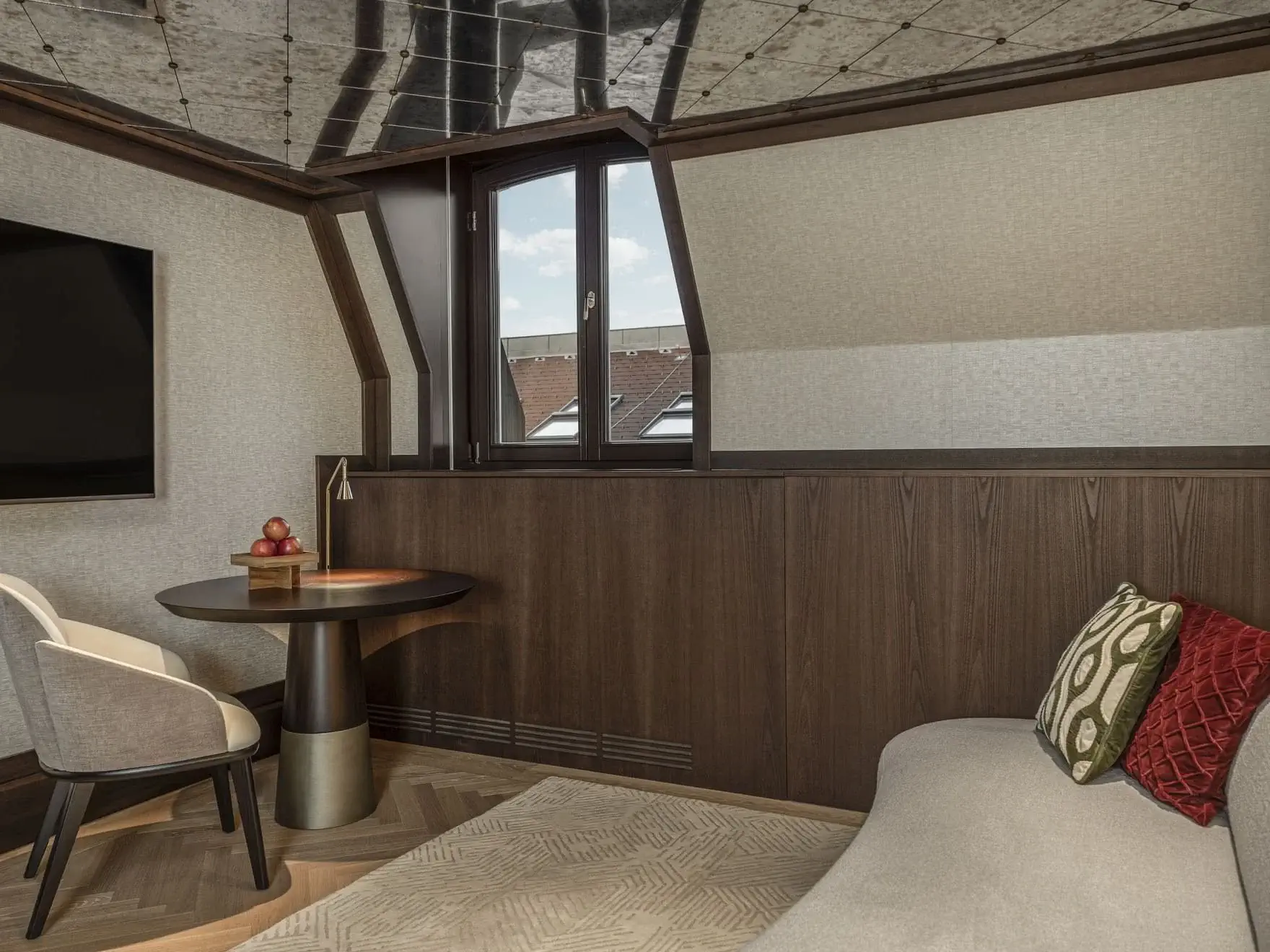 Modern attic room in the Almanac Suite with a small round table, two chairs, a flat-screen TV, a window, and a beige sofa with decorative pillows. Dark wood paneling and a patterned ceiling add character.