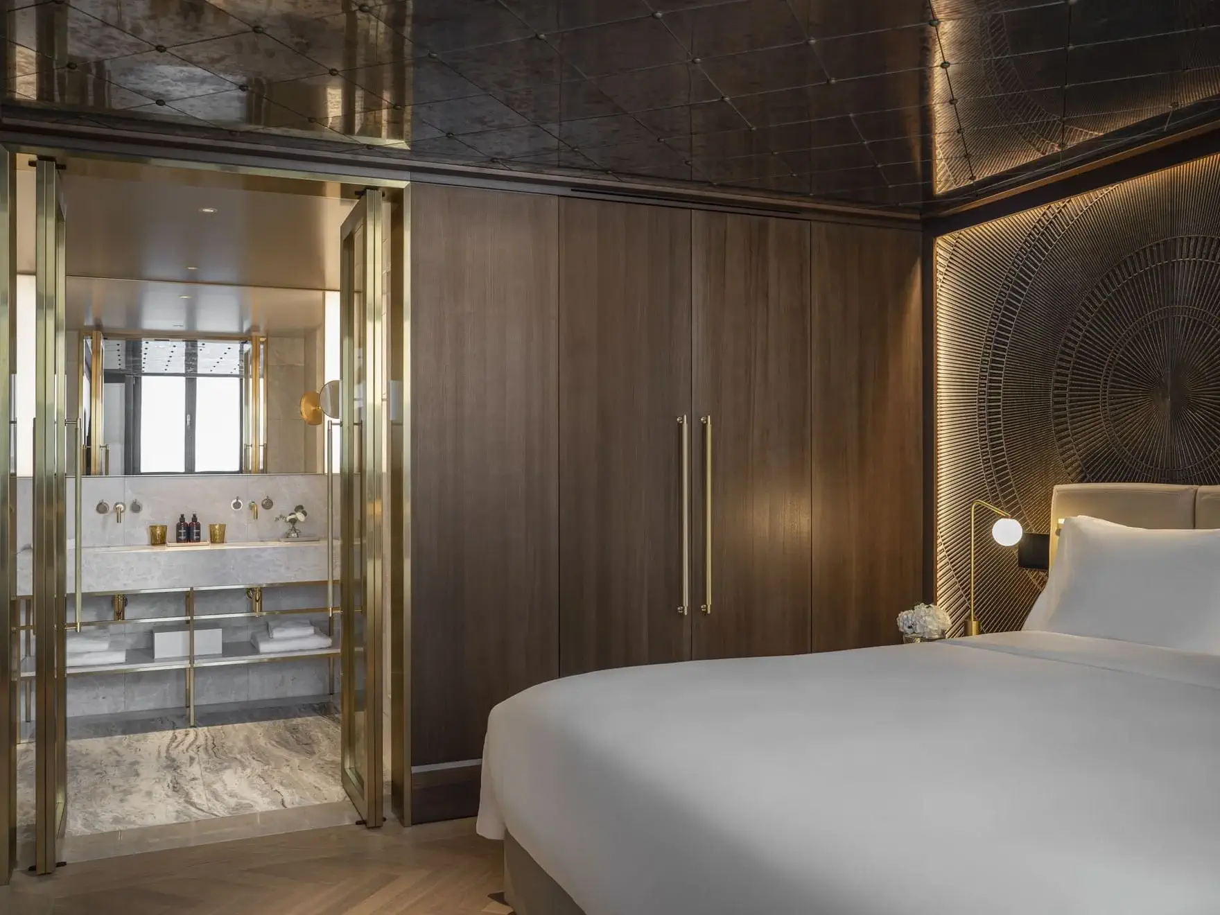 A modern Almanac Suite featuring a large bed, wood-paneled walls, decorative wall art, and a view into a marble bathroom with gold accents and glass doors.