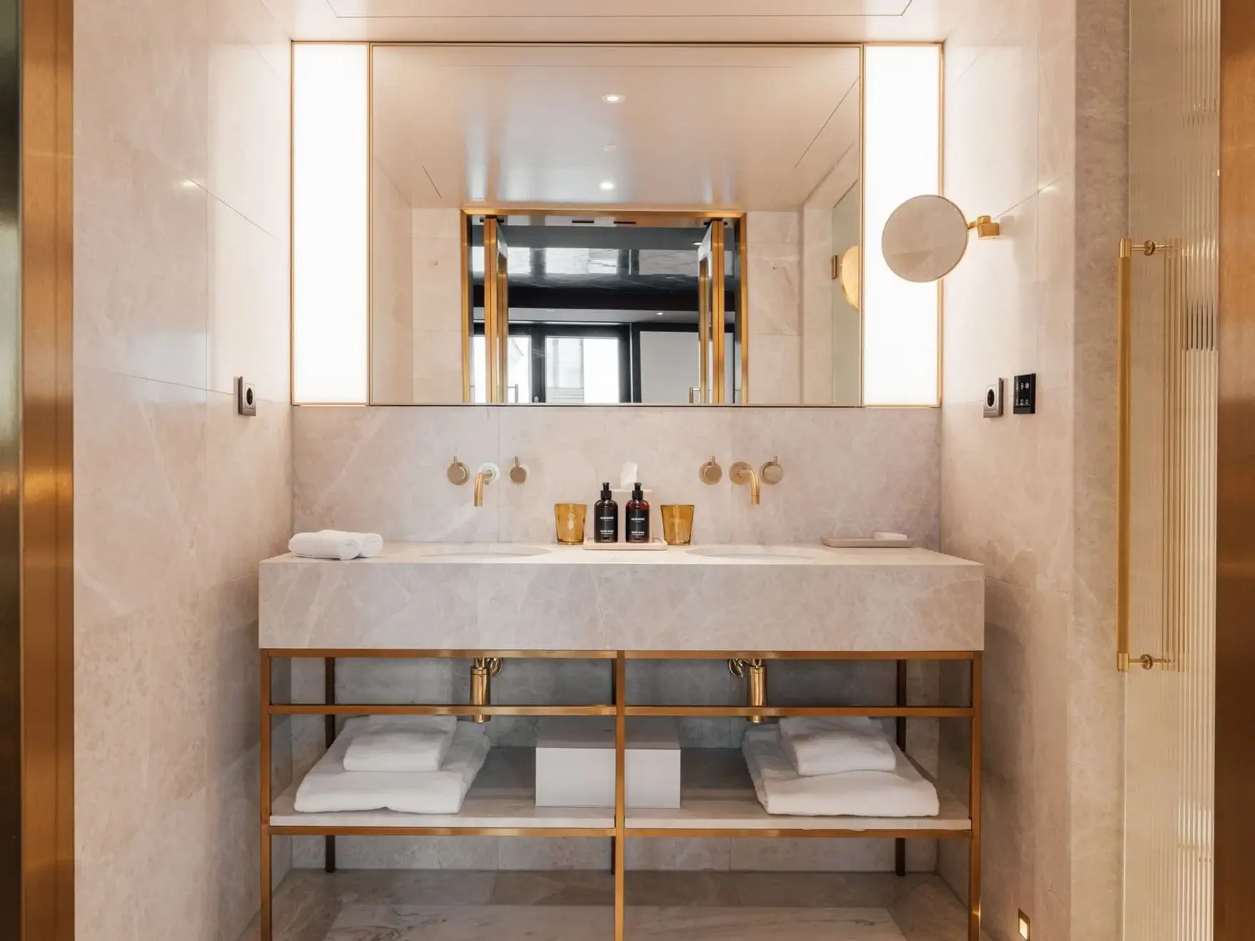 Modern bathroom in the Almanac Suite featuring a double sink vanity, gold fixtures, large mirror, under-sink shelving with towels, and elegant marble walls. Bottles and towels are thoughtfully placed on the countertop.