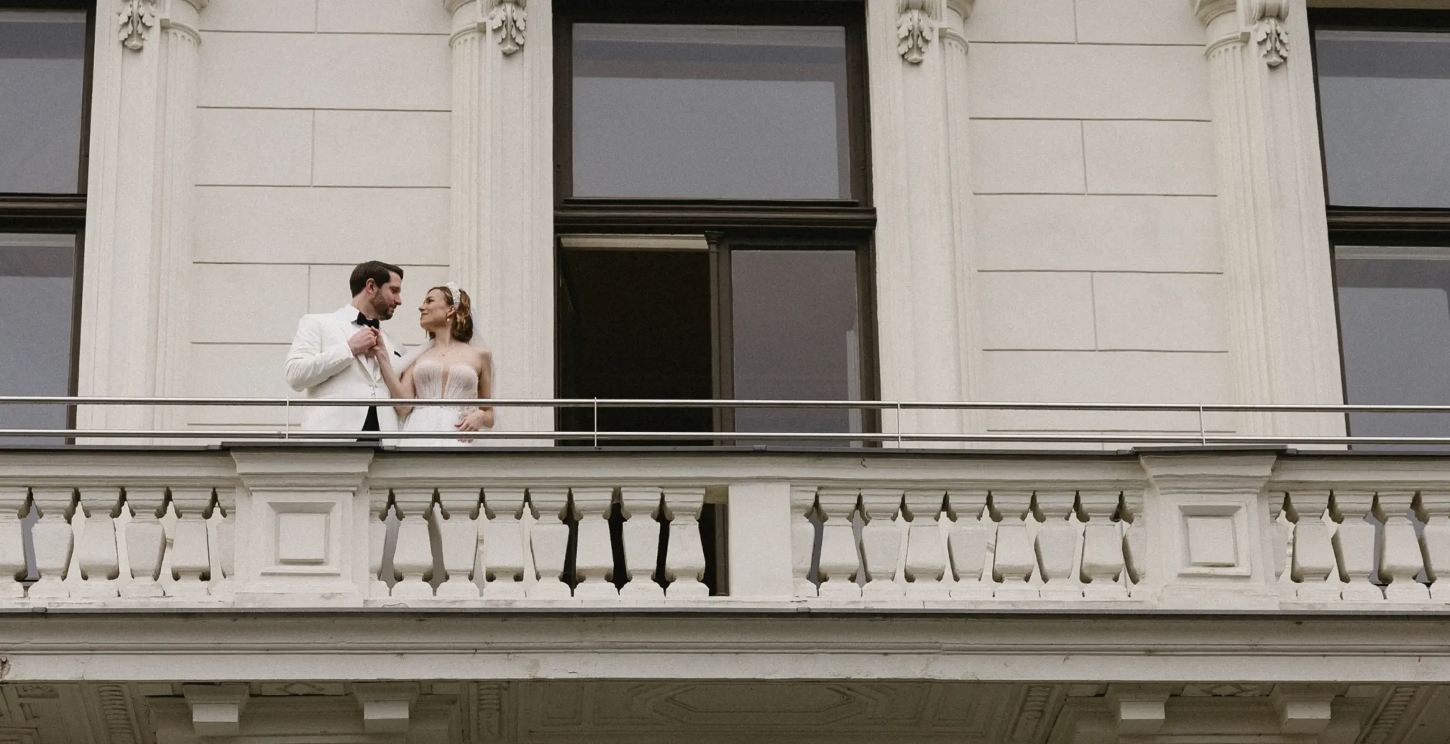 A bride and groom stand together on a white balcony of an ornate building, looking at each other.
