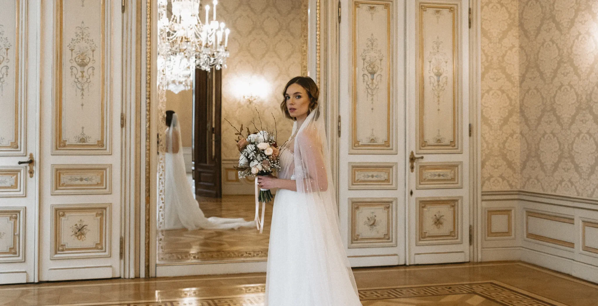 A bride in a white wedding dress holds a bouquet and stands in an ornate room with cream-colored walls, a large mirror, and chandelier.