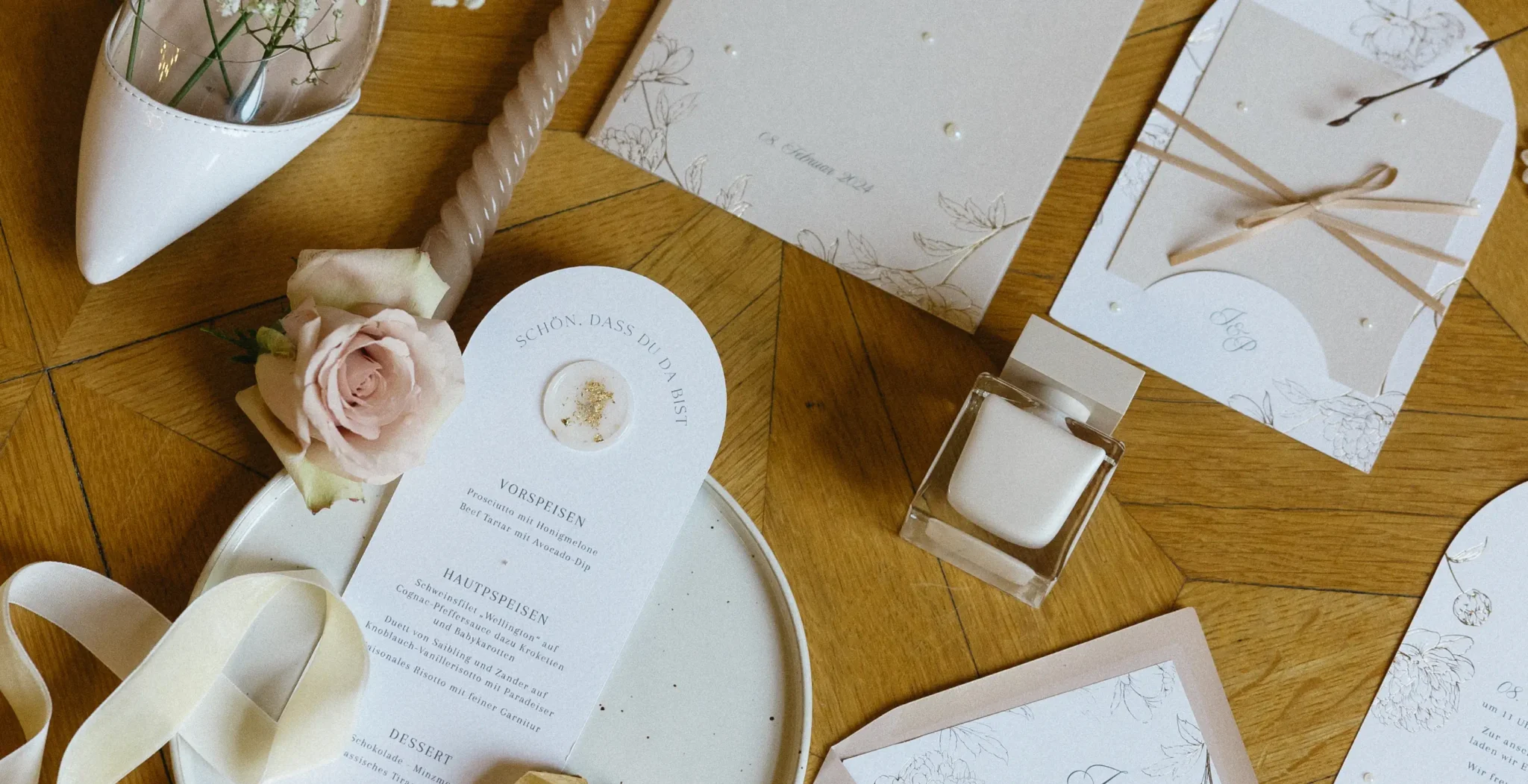 A flat lay of wedding stationery, including invitations, a menu, a candle, a pale pink rose, ribbon, and a small glass bottle, arranged on a wooden surface.