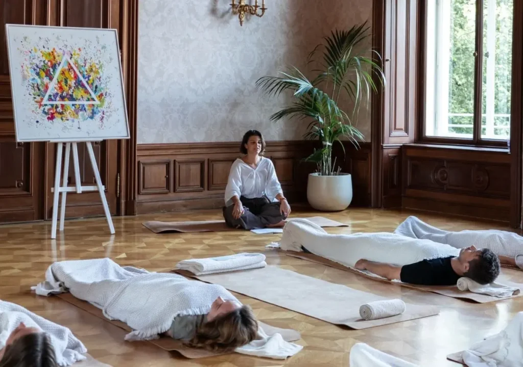 A person leads a relaxation or meditation session in a room where participants lie on mats covered with blankets; an abstract painting is displayed on an easel nearby.