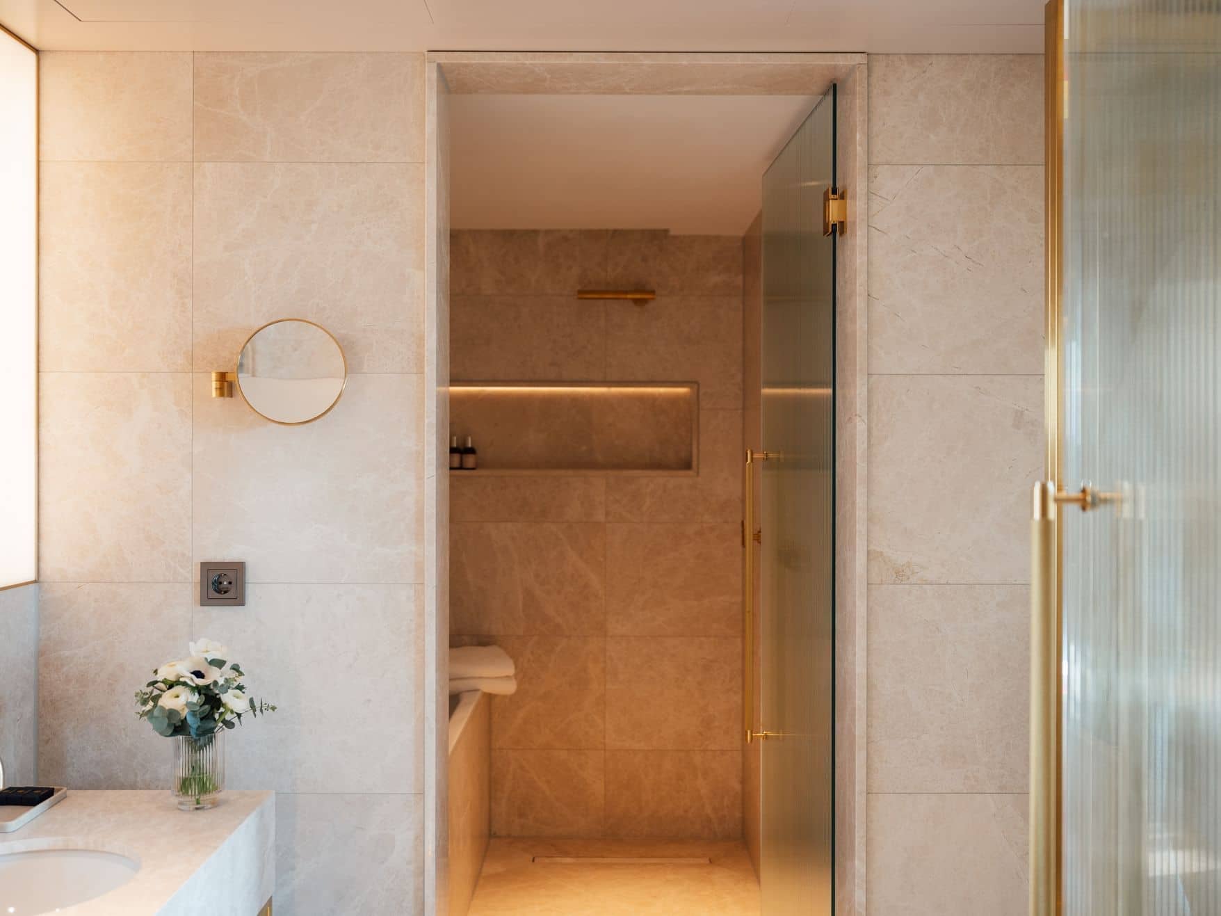 Modern bathroom in the Almanac Room with beige marble walls, a walk-in shower with glass door, a round mirror, and a small vase of flowers on the counter.