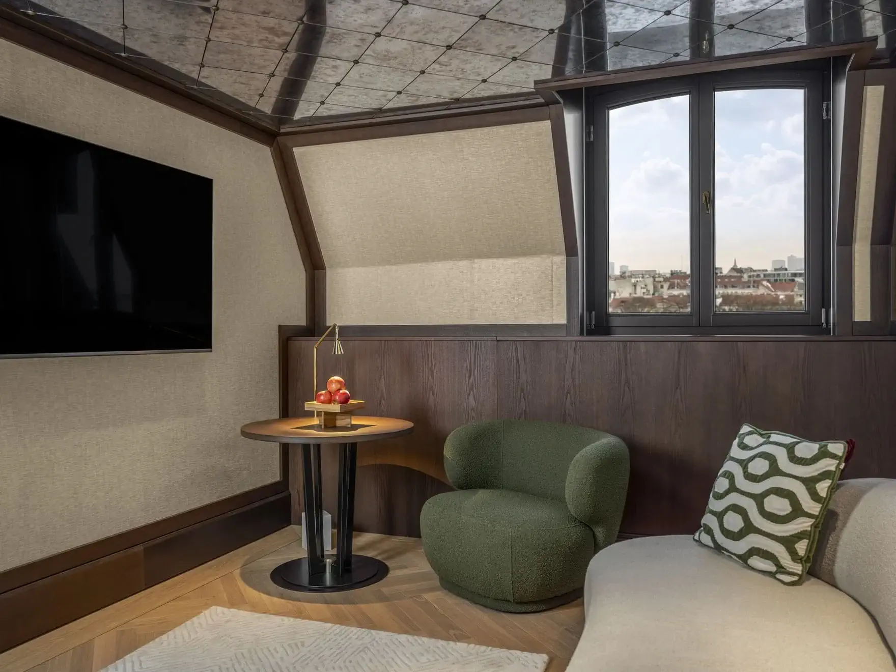 A modern Almanac Park Suite features a wall-mounted TV, a round table with a lamp and apples, a green chair, a beige sofa with a patterned pillow, and a window offering a city view.