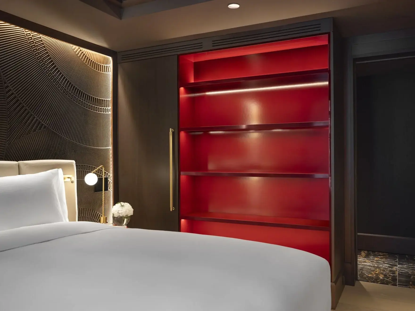 A Family Room with a neatly made bed, dark walls, a bedside lamp, and an empty red-lit shelving unit built into the wall.