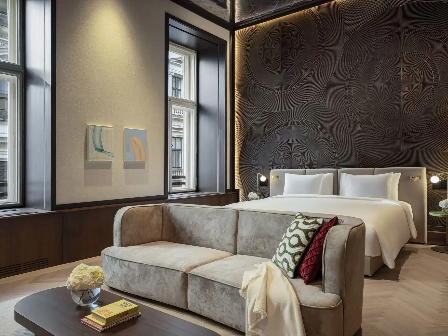 Modern Art Suite in Vienna featuring a double bed, beige sofa, coffee table with books and flowers, large windows, and abstract wall art above the bed.