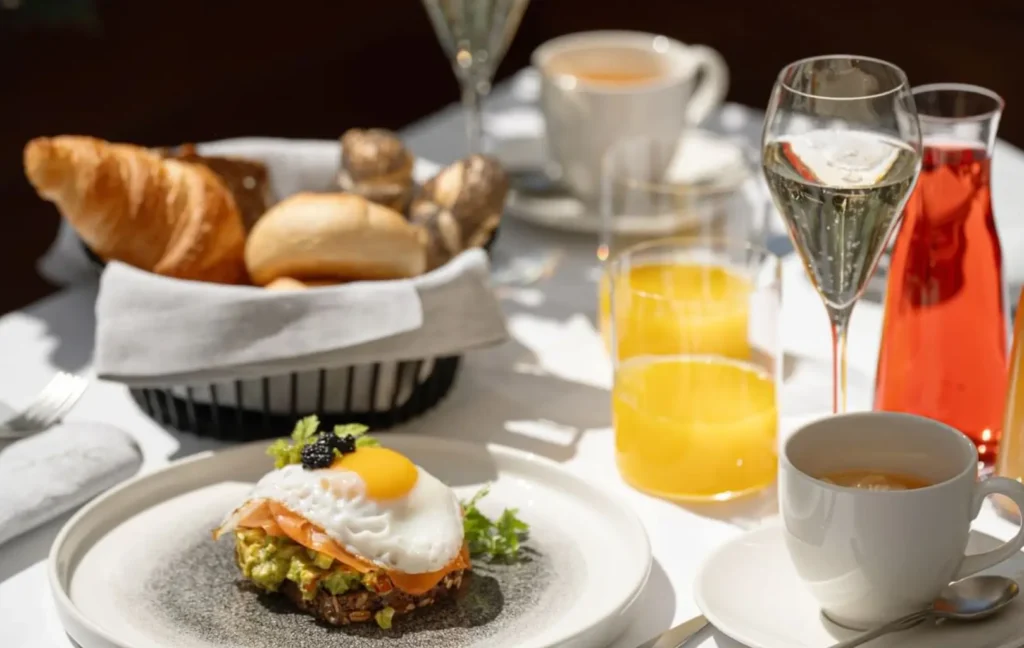 Beginnen Sie Ihren Tag mit Frühstück Mit Uns: ein Frühstückstisch mit Toast, belegt mit Avocado, Lachs und einem Spiegelei, dazu ein Korb mit verschiedenen Brotsorten, Saft, Kaffee und prickelnden Getränken.