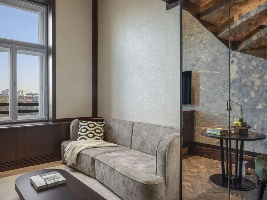 Modernes Duplex-Wohnzimmer mit einem beigen Sofa, einem gemusterten Kissen, einer Decke, einem Couchtisch aus Holz und einem großen Fenster mit Blick auf die Stadt. Eine Glaswand trennt den Essbereich in dieser stilvollen Park Suite ab.
