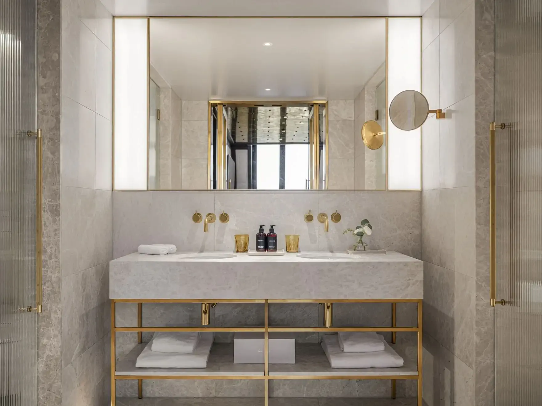 Modern Duplex Park Suite bathroom with double sinks, marble countertop, gold fixtures, large mirror, toiletries, rolled towels, and shelves below holding additional towels.