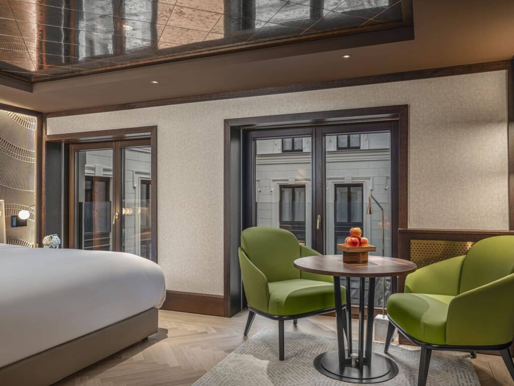 Modernes Almanac-Hotelzimmer mit einem Bett, zwei grünen Sesseln, einem kleinen runden Tisch mit Obst, großen Fenstern und neutralem Dekor.