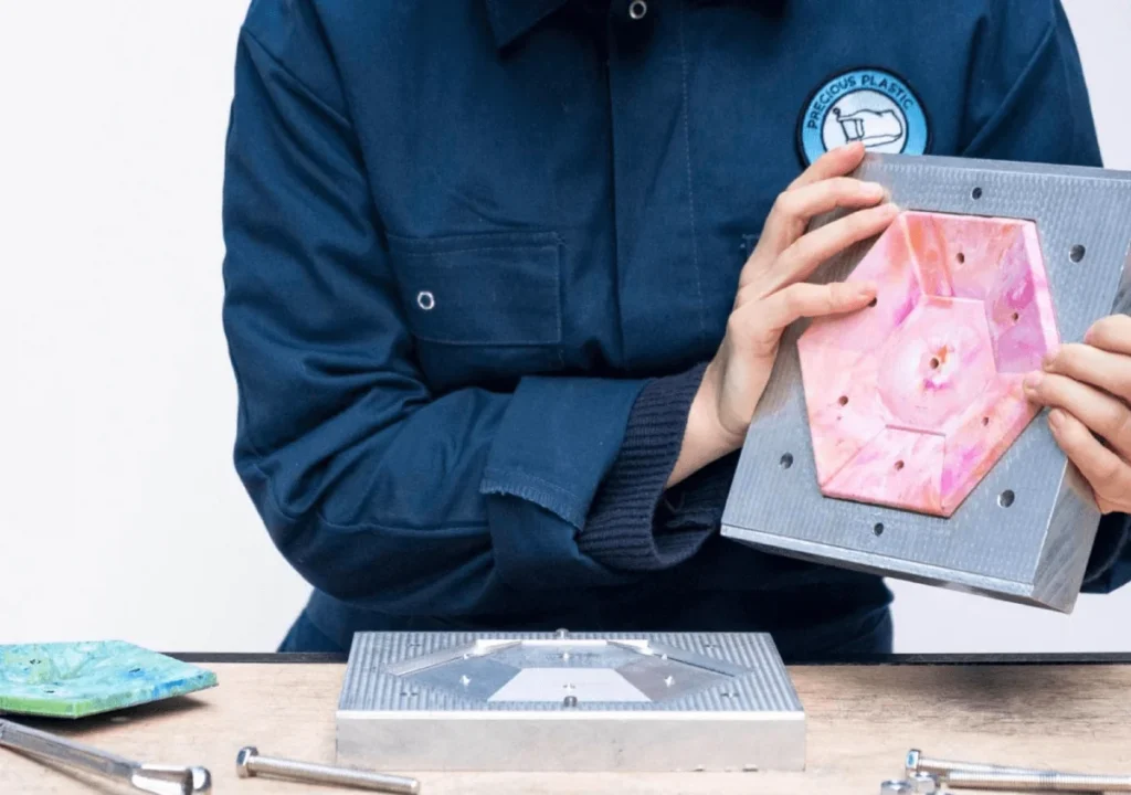 Person in blue coveralls holding a hexagonal pink plastic piece placed in a metal mold on a table with tools and another mold piece nearby, demonstrating careful attention to social responsibilities in manufacturing.