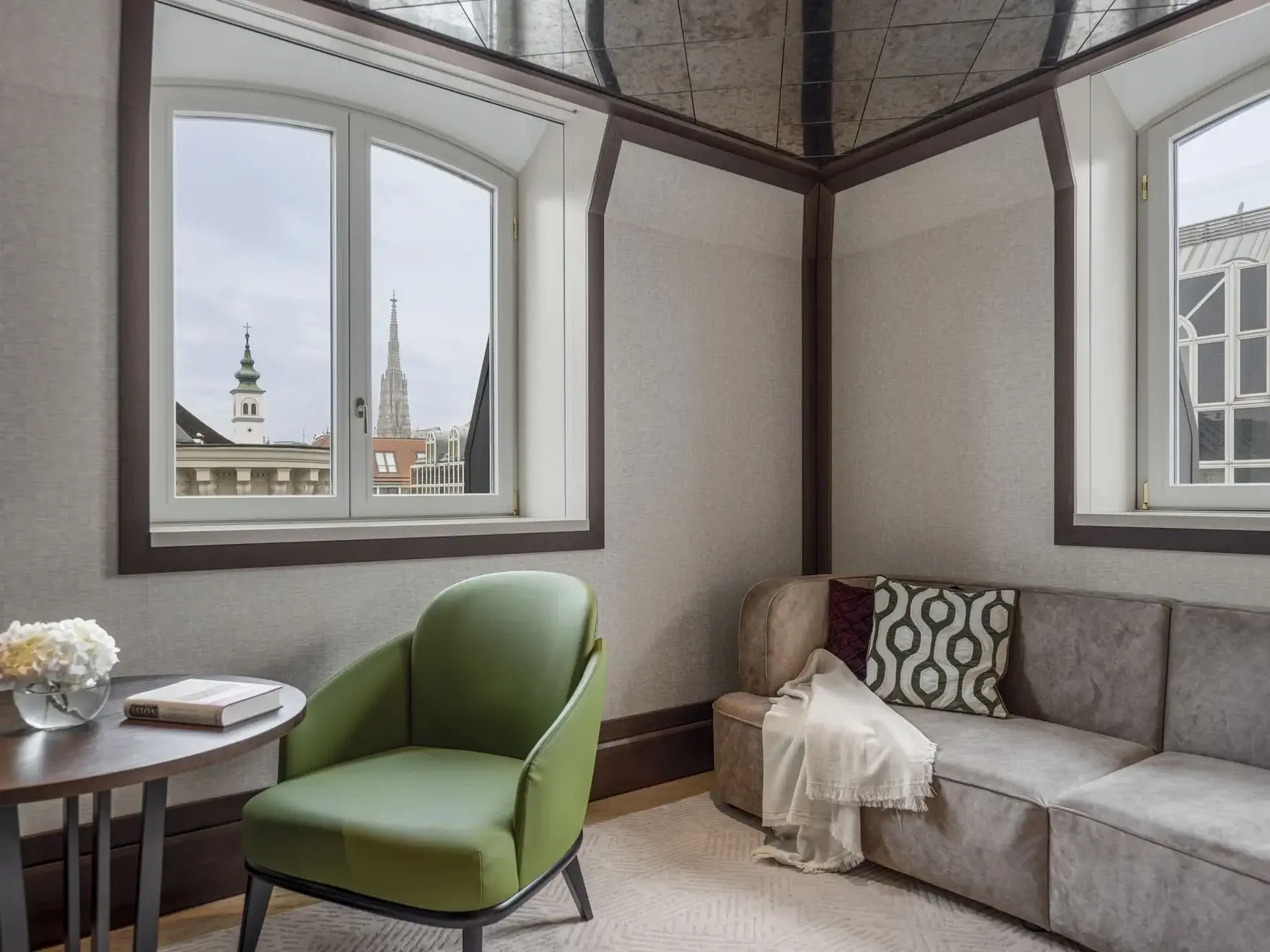Modern lounge area with green chair, round table, and L-shaped sofa near windows overlooking a cityscape with church spires, exuding the refined elegance of a Palais Suite.