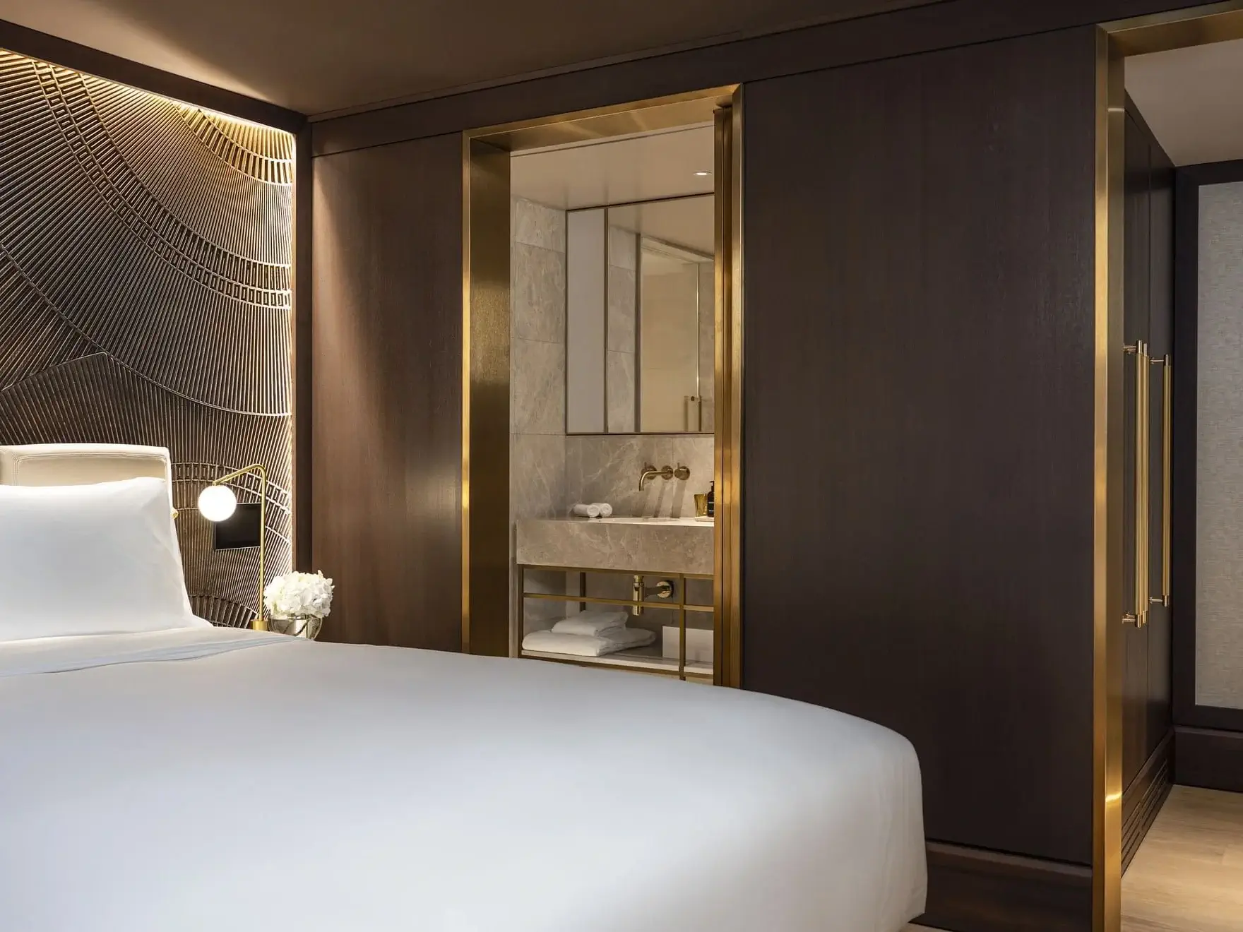 A modern Family Two Bedroom hotel room with a neatly made bed, dark wood paneling, gold accents, and a view into a marble bathroom with a sink and towels.
