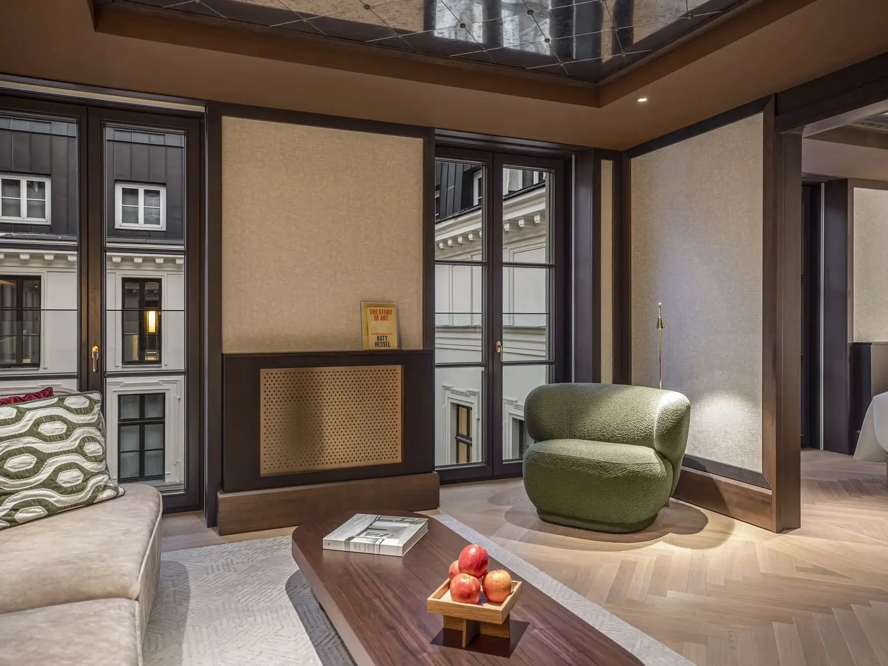 Modern Family Two Bedroom living room with large windows, green armchair, light sofa, wooden coffee table with books and a bowl of apples, and neutral-toned walls. Urban building visible outside.