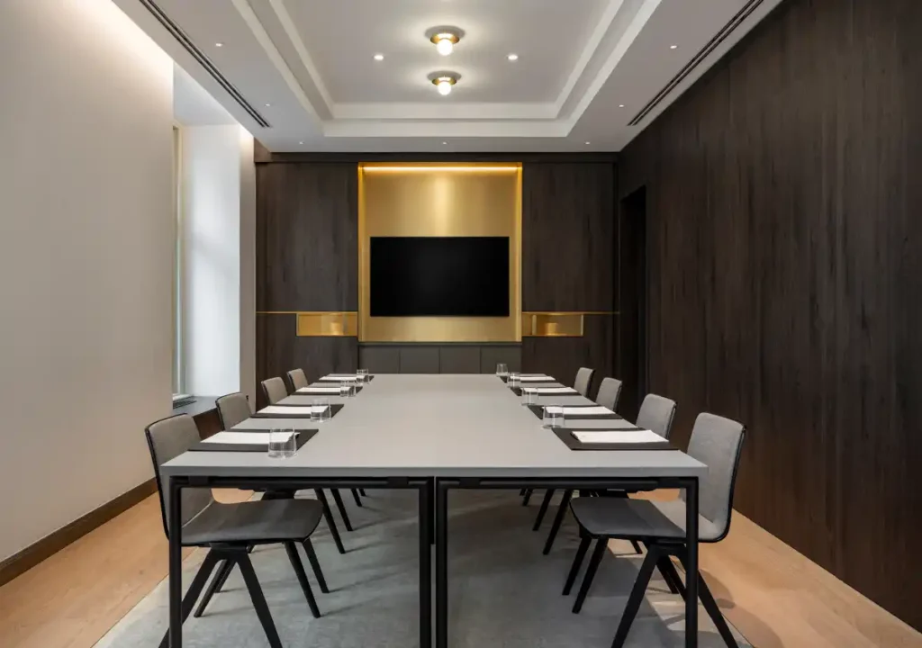 Modern conference room ideal for meetings or events, featuring a rectangular table, ten chairs, notepads, glasses of water, a wall-mounted TV, and wood-paneled walls with accent lighting.
