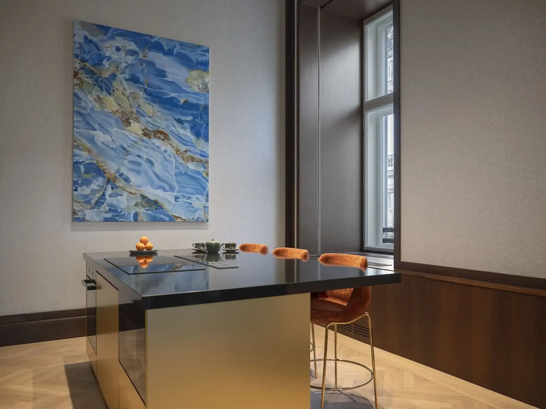 A modern kitchen in the Vienna Art Suite features a reflective gold island, three orange stools, a blue abstract painting on the wall, and large windows letting in natural light.