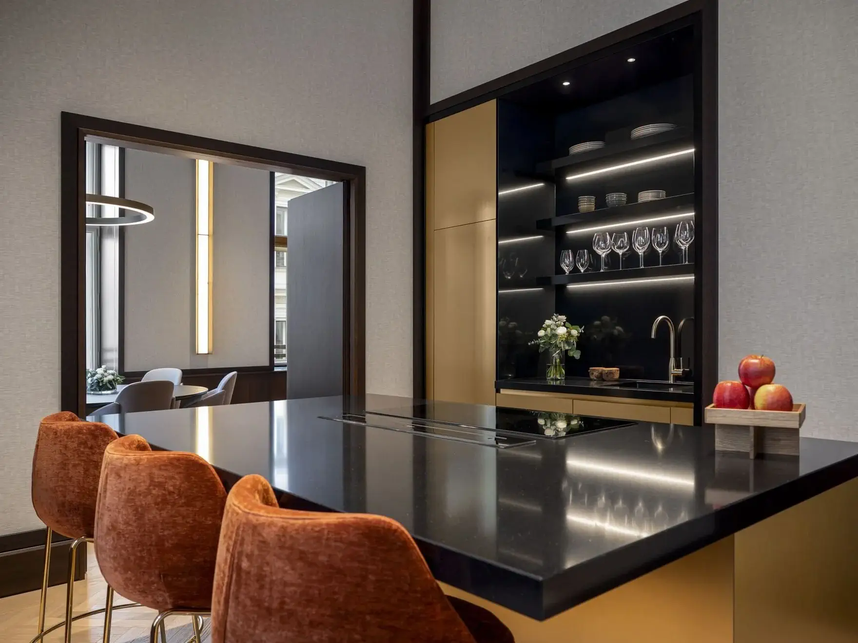 Modern kitchen in this Art Suite features black countertops, gold trim, rust-colored bar stools, open shelving with glassware, and a tray of apples. Dining area is visible through the doorway—bringing Vienna style into your home.