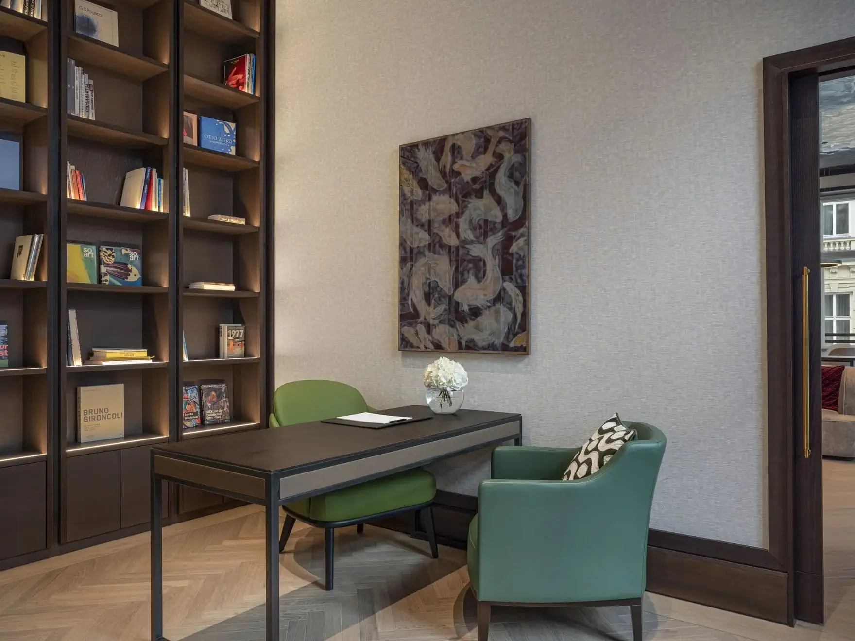 A modern office in Vienna with a dark desk, two green chairs, a vase of flowers, a large bookshelf filled with books, and abstract art on the wall creates an inspiring Art Suite atmosphere.