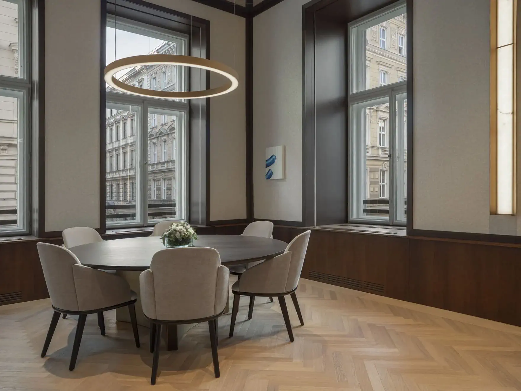 A modern Vienna conference room with a round table, six upholstered chairs, a circular ceiling light, large windows, and light wood herringbone flooring evokes the elegance of an Art Suite.