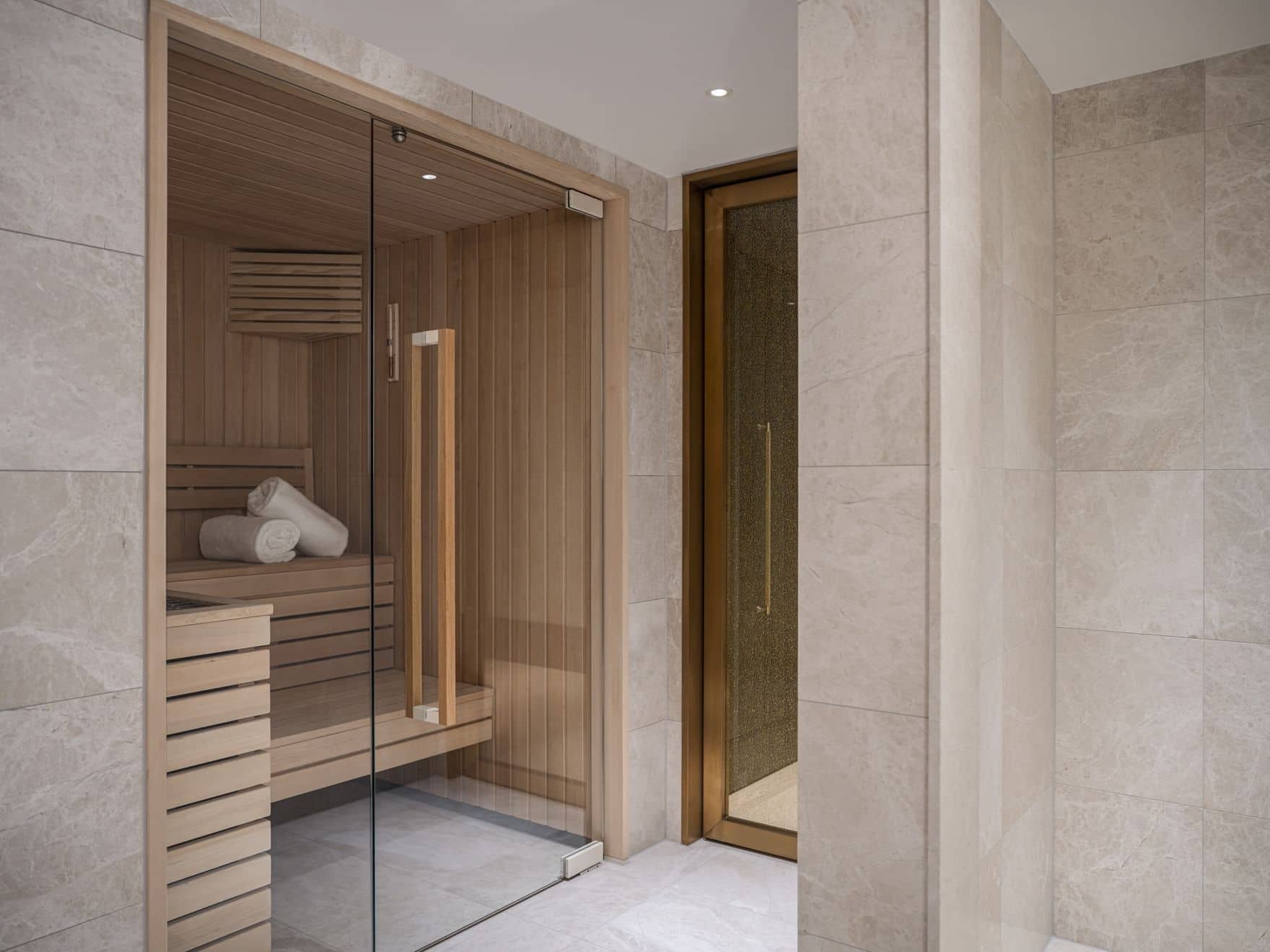 Modern spa area with a glass-enclosed wooden sauna, neatly rolled towels on a bench, and elegant Palais Floor tiles in neutral tones.