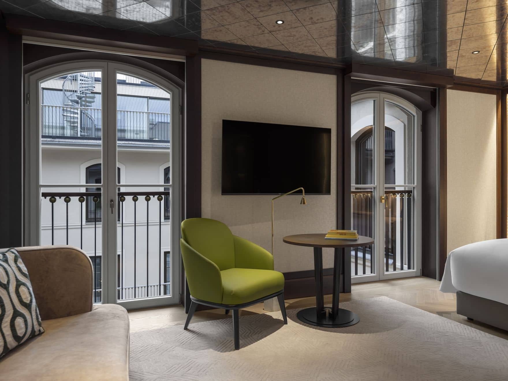 Modern hotel room with large arched windows, a wall-mounted TV, a green chair by a round table, and a partial view of bed and sofa. Enjoy balcony railings and the building exterior from this elegant Palais Floor retreat.