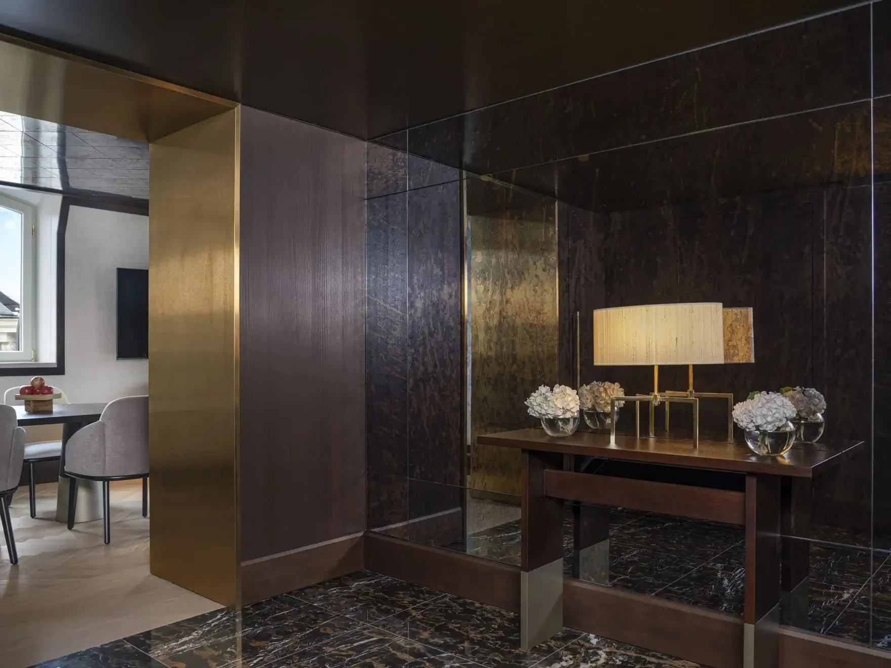 A modern interior in the Palais Suite features dark marble floors, a mirrored wall, and a console table with a lamp and vases of white flowers. Through an open doorway, a dining area with light chairs is visible.