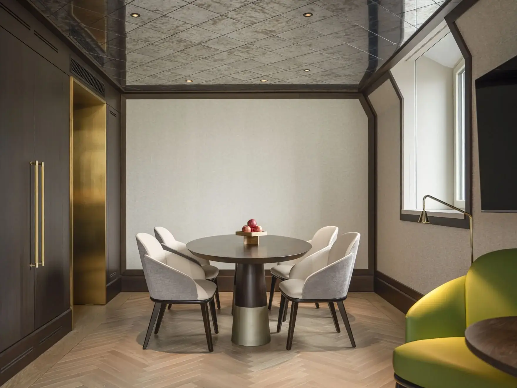 A modern meeting room in the Palais Suite features a round table, five upholstered chairs, a bowl of apples, wooden walls, large windows, and a green sofa in the corner.