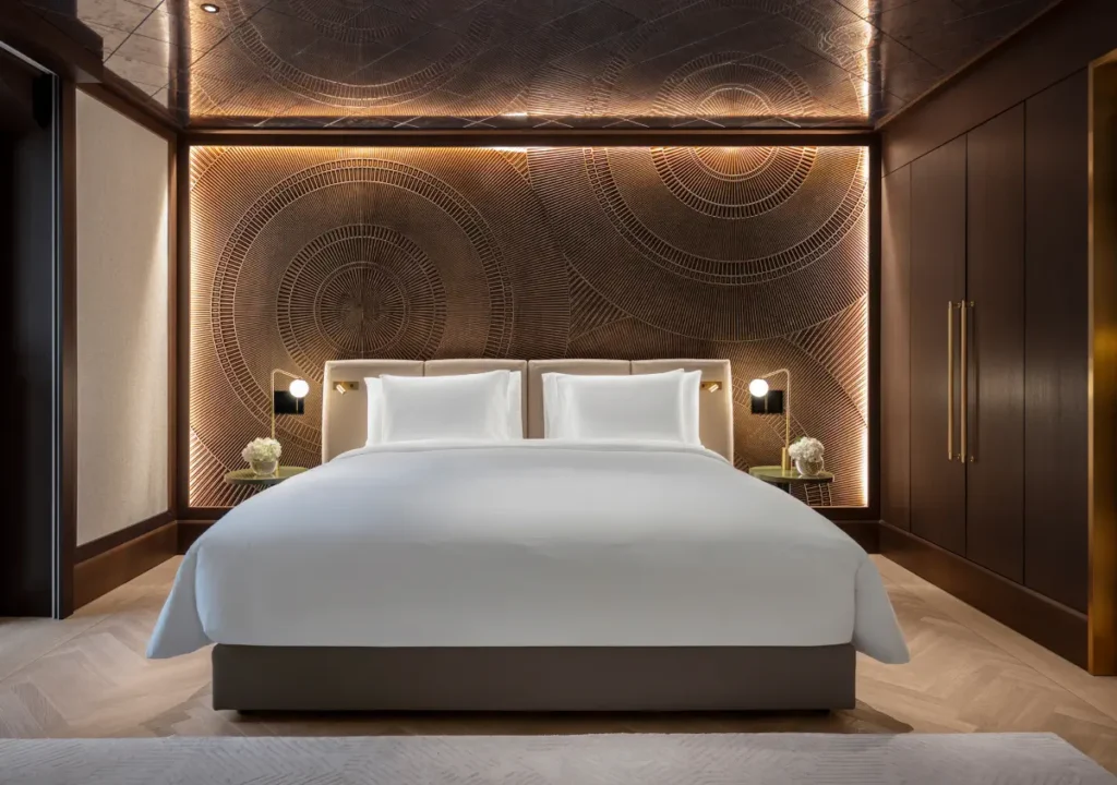 Modern hotel bedroom inspired by Palais Vienna, featuring a large white bed, two bedside tables with lamps and flowers, and decorative wall art behind the bed. Warm lighting and dark wood accents throughout create an inviting Almanac atmosphere.