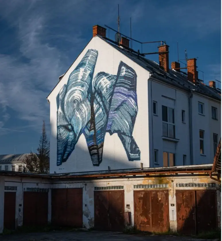 A large abstract mural with blue, black, and purple tones is painted on the side of a white building with several chimneys, above a row of old garages.