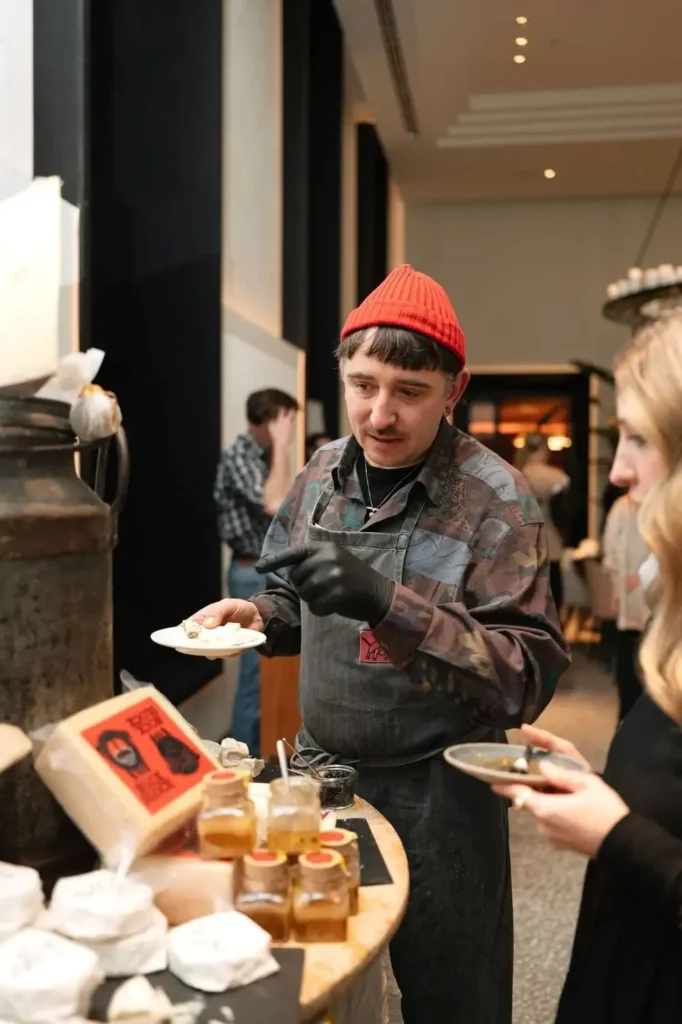 A man in a red beanie and gloves serves samples of cheese and spreads to a woman at an indoor Donnersmarkt Restaurant market stall.
