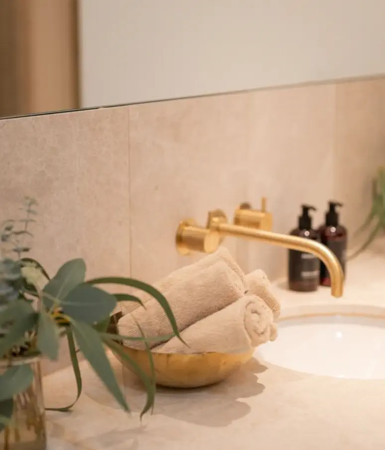 Rolled beige towels in a gold bowl on a bathroom counter with a gold faucet, two soap dispensers, and green plant stems add elegant charm reminiscent of Almanac Palais Vienna.
