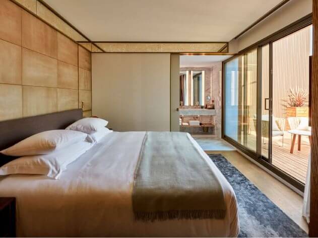 Eine moderne Terrassensuite mit einem Kingsize-Bett mit weißer Bettwäsche und grüner Decke, angrenzend an ein Badezimmer und eine private Terrasse, die durch große Glasschiebetüren zu sehen ist.
