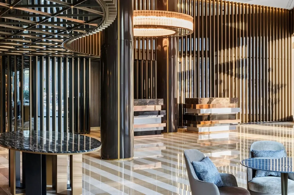 Moderno vestíbulo de hotel con suelos de mármol a rayas, pilares metálicos, techo geométrico, lámpara de araña circular y zona de asientos con sillas grises y mesas de mármol.