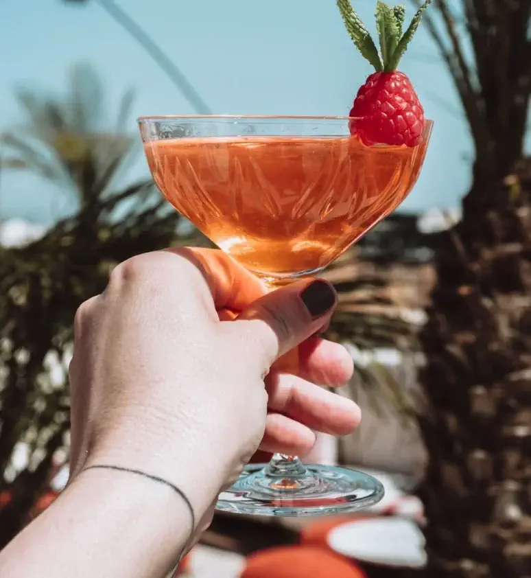 A hand holding a coupe glass filled with an amber cocktail, garnished with a raspberry and a mint sprig, with palm trees in the blurred background.