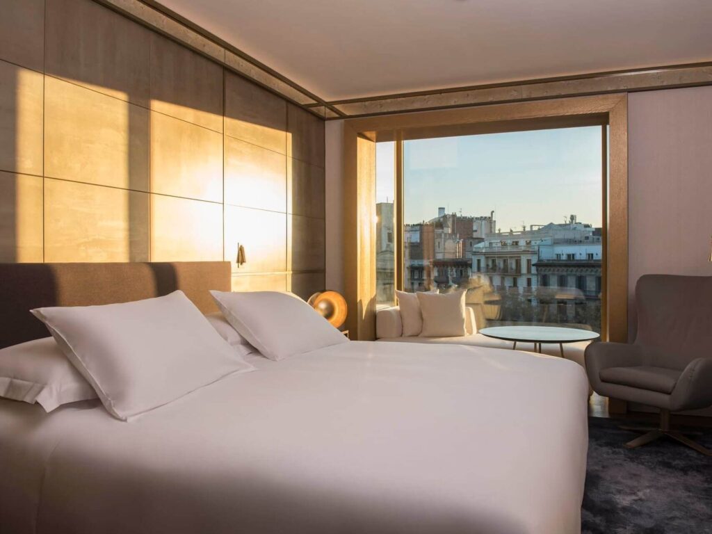 Moderne Almanac Suite mit großem Bett, weißer Bettwäsche, Sessel, kleinem Tisch und bodentiefem Fenster mit Blick auf eine Stadtlandschaft bei Sonnenuntergang.