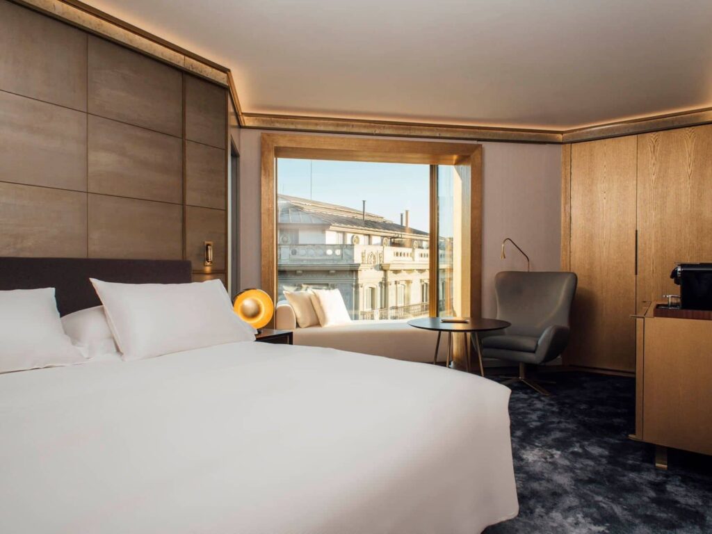 Una moderna Habitación Almanaque con una cama grande, un sillón junto a una mesa redonda, paredes con paneles de madera y una ventana con vistas a los edificios de la ciudad.