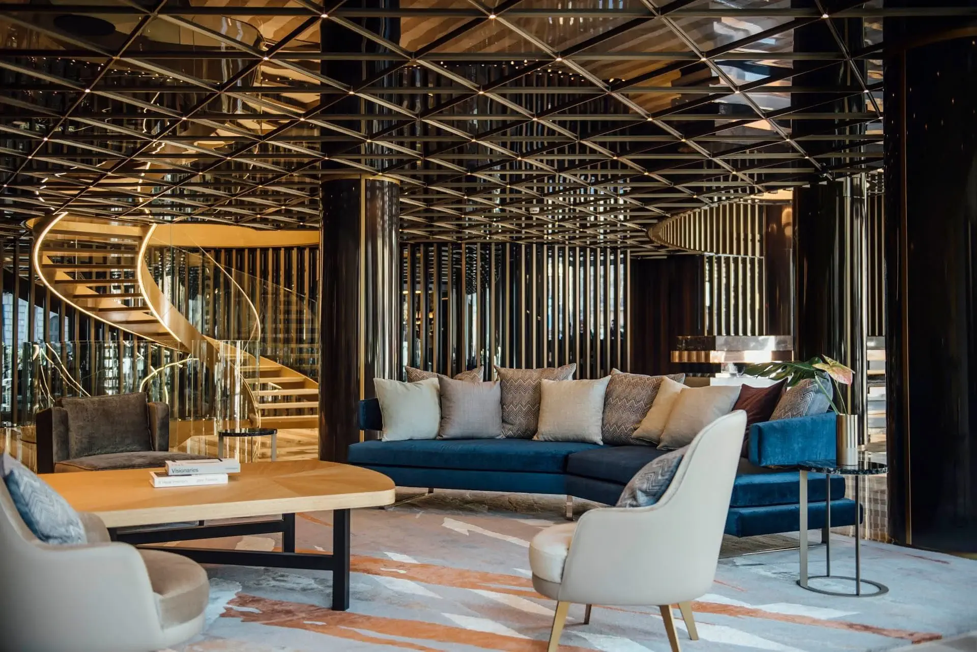 Moderno vestíbulo de hotel con asientos azules y beige, una mesa de centro de madera, una escalera de caracol y un techo decorativo de espejos con motivos geométricos.