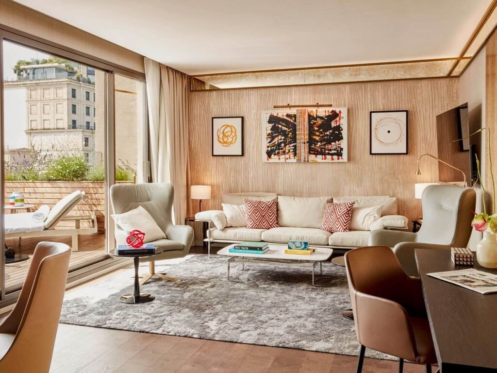 Modernes Wohnzimmer in einer Grand Penthouse Suite mit beigefarbenem Sofa, Sesseln und Wandkunst, mit großen Fenstern, die auf einen Balkon mit Sitzgelegenheiten im Freien und Stadtblick führen.