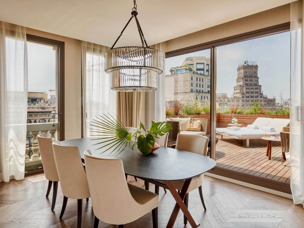 Moderner Essbereich in einer Penthouse Suite mit sechs weißen Stühlen um einen dunklen ovalen Tisch, großen Fenstern, Blick auf die Stadt, Terrasse mit Sitzgelegenheiten im Freien, Grünflächen und Zugang zu zwei Schlafzimmern.