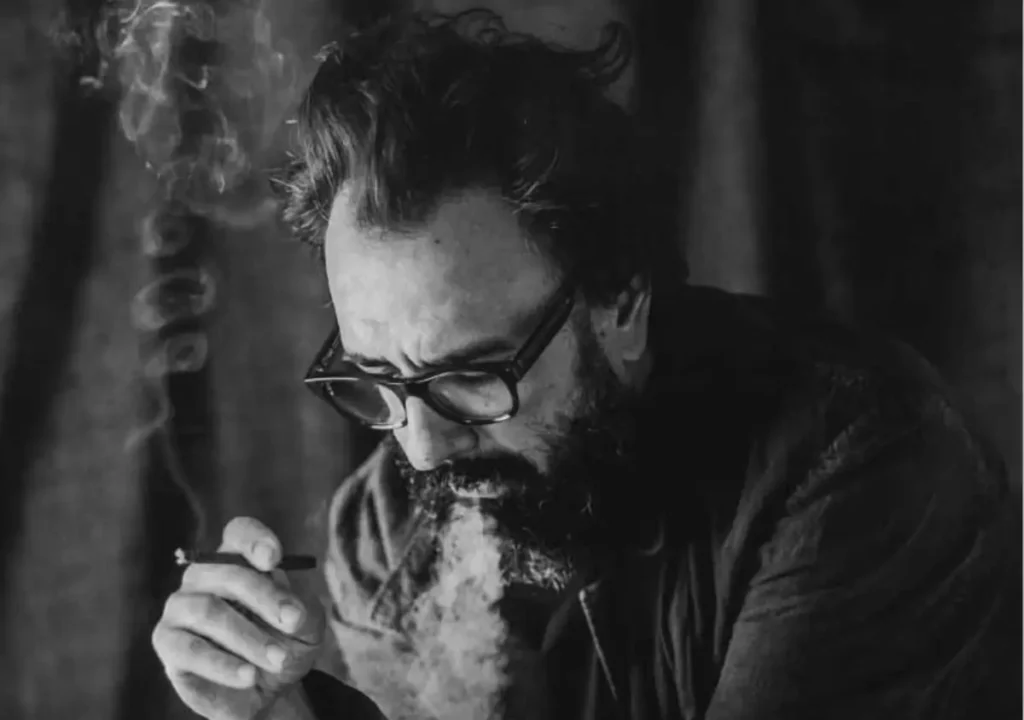 Una persona con barba y gafas fuma un cigarrillo, con humo visible rodeando su cara en un entorno poco iluminado.