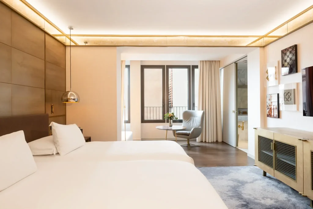 Modernes Hotelzimmer mit zwei Doppelbetten, einem Stuhl mit kleinem Tisch, Wandmalerei, großem Fenster und offenem Bad mit Glasduschtür.