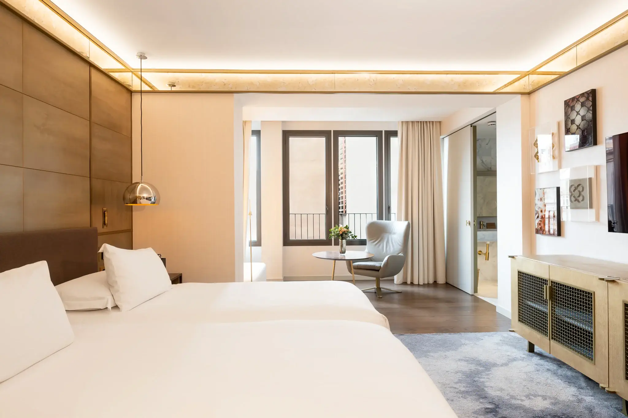 Modernes Hotelzimmer mit zwei Betten, einem Stuhl am Fenster, hellem Holzfußboden, zeitgenössischem Dekor und einem durch eine Glastür einsehbaren Bad.