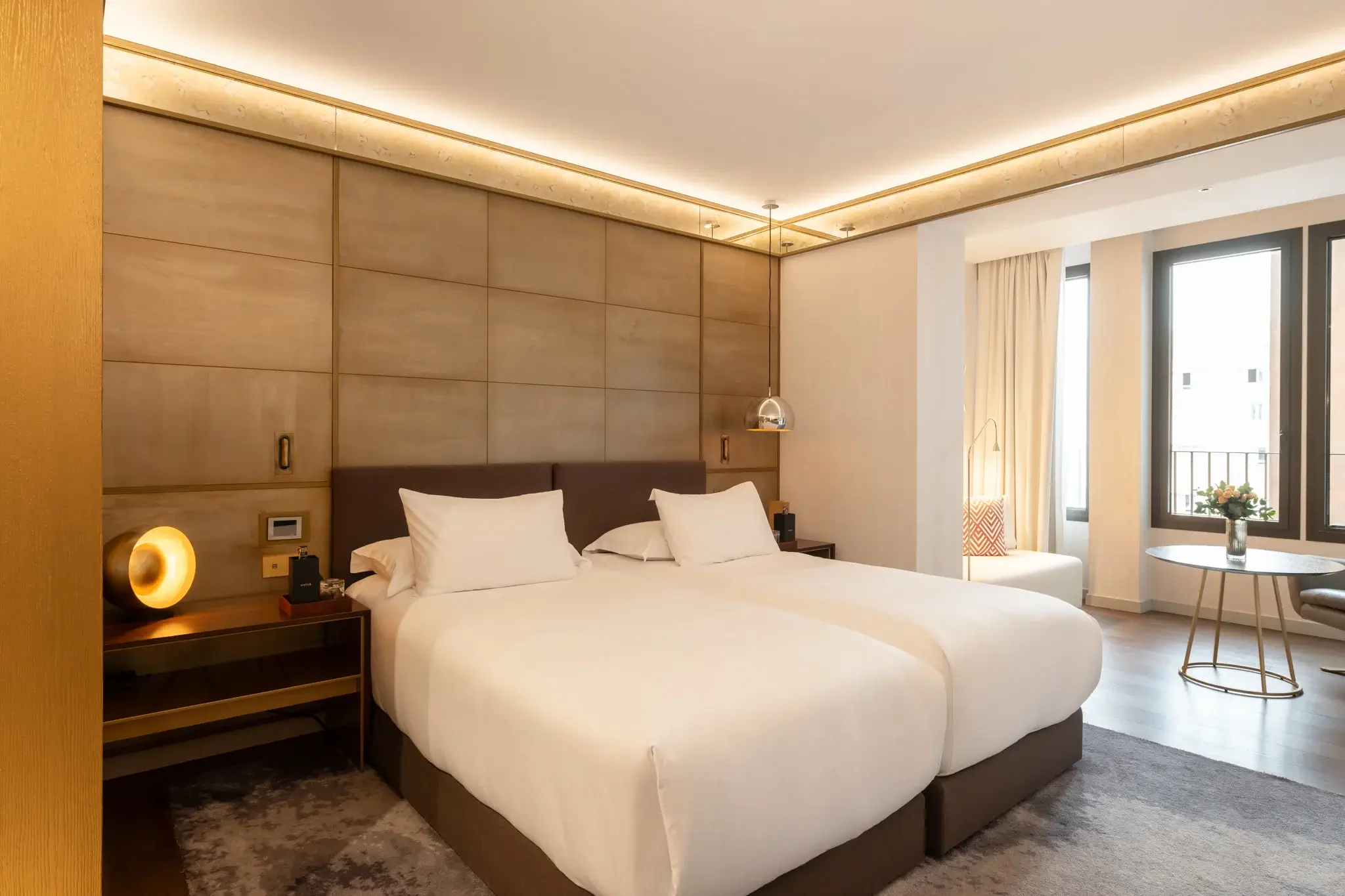 Modernes Hotelzimmer mit zwei Einzelbetten, einem Kopfteil aus Holz, Nachttischen, einem runden Tisch mit Stühlen in der Nähe der großen Fenster und einem Dekor in neutralen Farben.