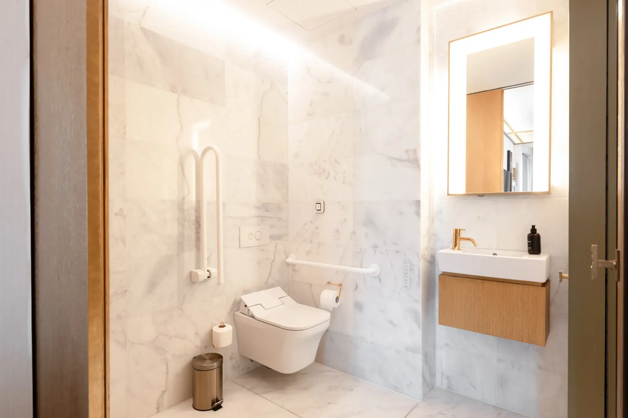 Ein modernes, barrierefreies Badezimmer mit Marmorwänden, einer wandmontierten Toilette mit Stützstangen, einem schwimmenden Waschbecken, einem Spiegel und einem Mülleimer.