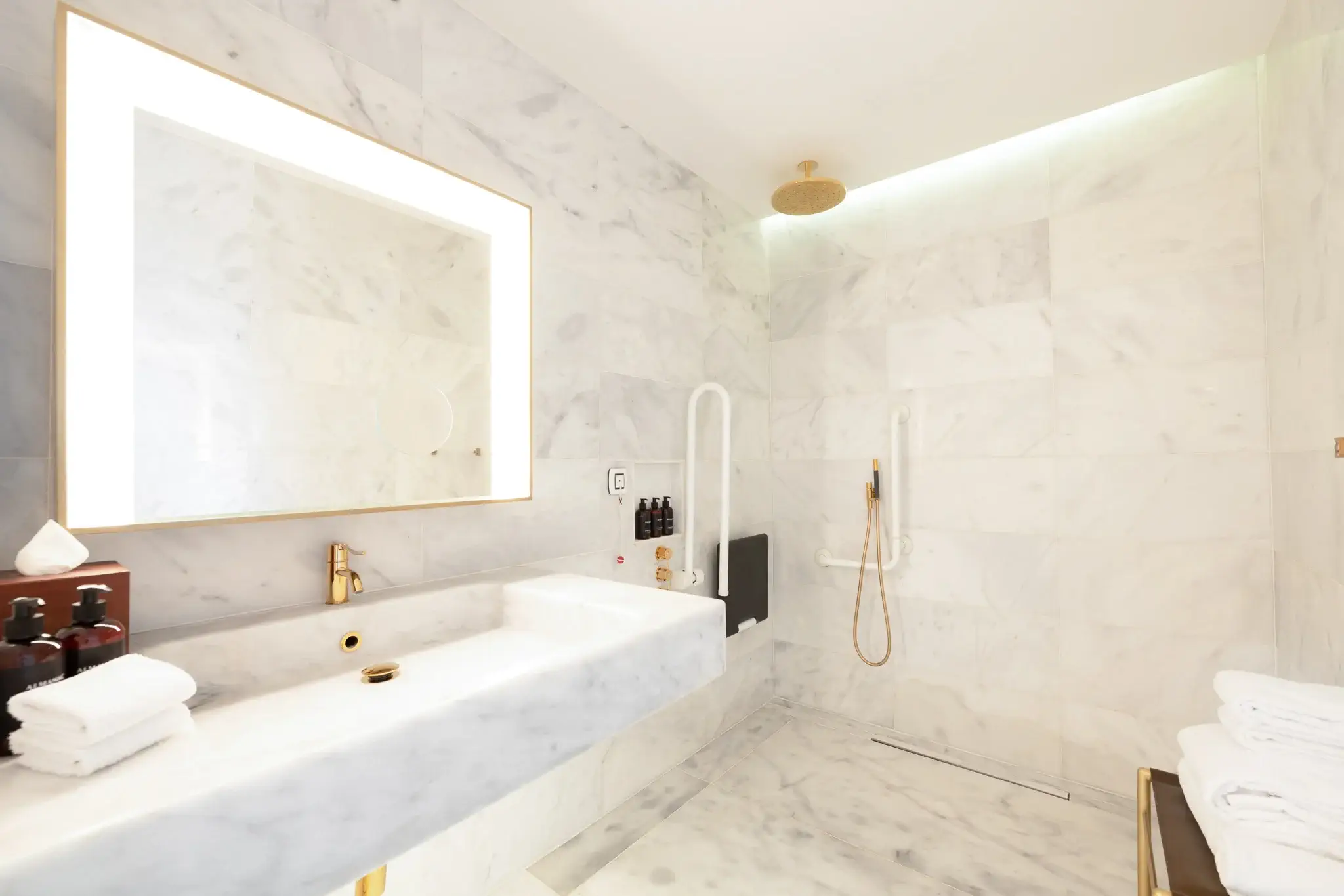 Ein modernes, behindertengerechtes Badezimmer mit Marmorwänden, einem großen, beleuchteten Spiegel, einer rollbaren Dusche, einem klappbaren Sitz und goldenen Armaturen. Handtücher und Toilettenartikel sind übersichtlich angeordnet.